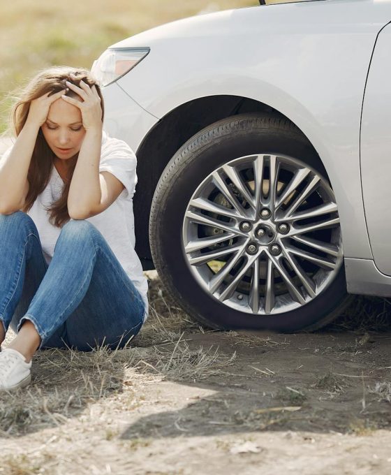 Frustrated female driver in white t shirt and jeans sitting on ground near damaged car with hands on head during car travel in sunny summer day